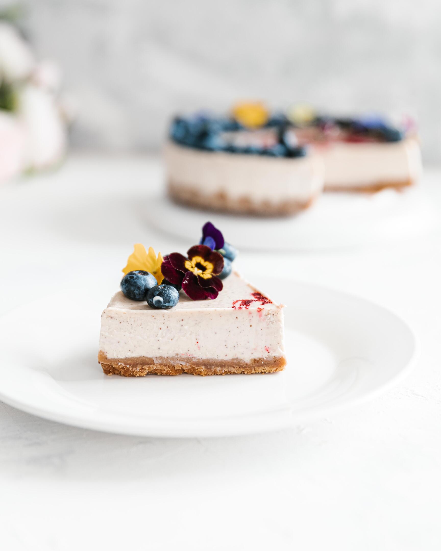 A piece of cheesecake is on a plate.  A piece of cheesecake is on a plate. It is decorated with edible flowers and blueberries. There is a cheesecake in the background.