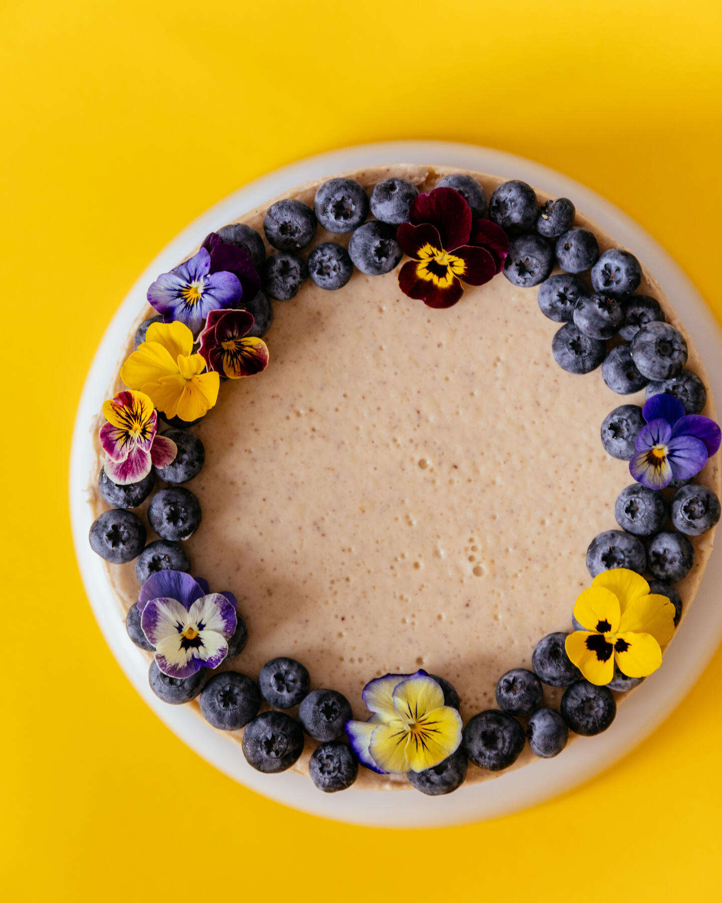 Cheesecake stands on a yellow background. Cheesecake stands on a yellow background. Photo from above. It is decorated with edible flowers and blueberries.