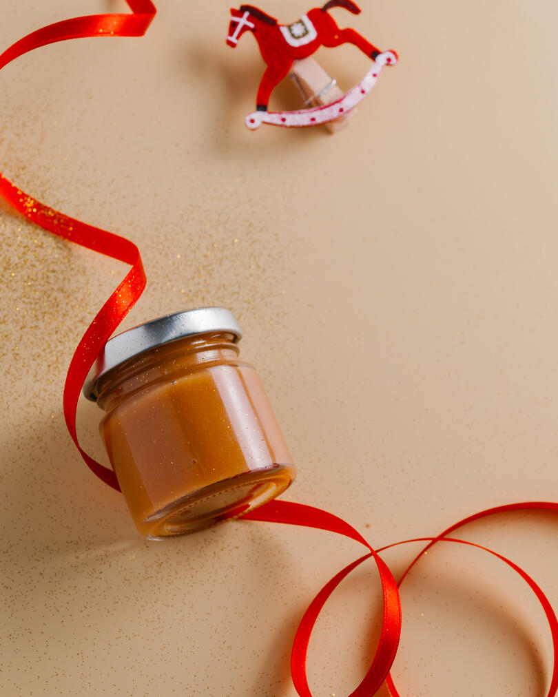 Home made caramel in small jar. On a beige background lies a jar of caramel. A red satin ribbon is wrapped around the jar and sequins are scattered.
To the right of the jar, a little above is a clothespin in the shape of a red horse. The photog with its colors reminds the viewer of the New Year's mood.
