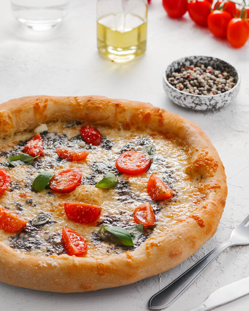 Пицата е на масата. На масата има ароматна пица. На заден план на рамката има домати и чаша вода.