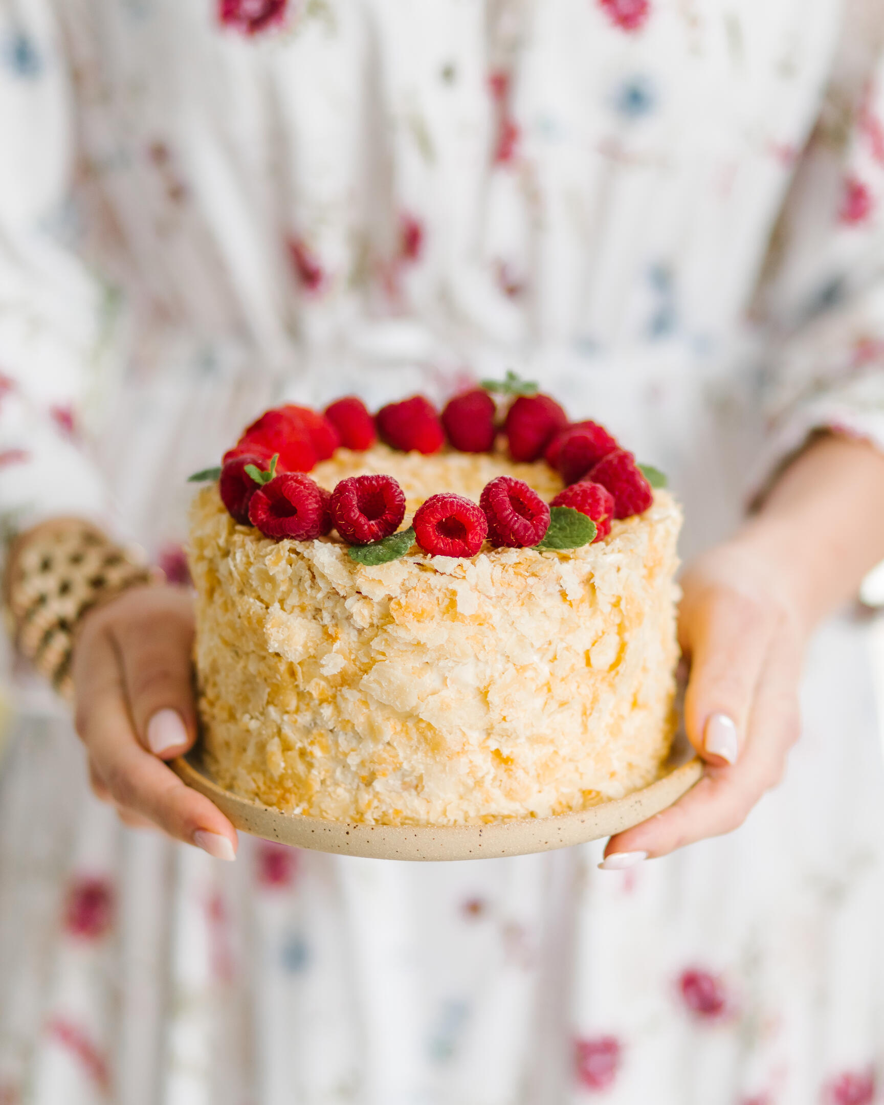 Жена държи чиния с торта в ръцете си.  Жена в лека рокля с флорален принт държи чиния с торта в ръцете си. Тортата се украсява с малини. Торта Наполеон.