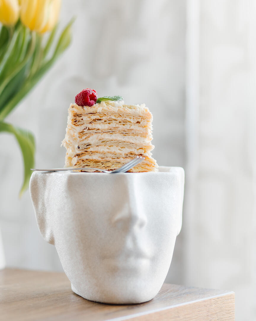 Торта Наполеон в чиния във формата на глава Торта Наполеон в чиния във формата на глава. На заден план има жълти лалета. До тортата има десертна вилица.