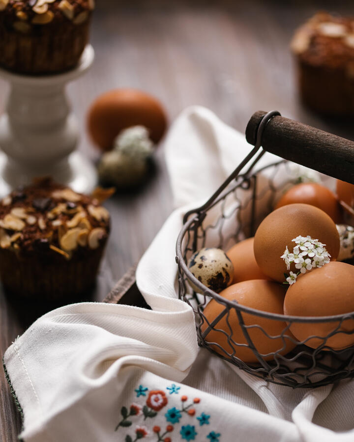 There is a carrot cake in the small basket.  Chicken eggs are in a metal basket. Under the basket is an embroidered towel. There is a carrot cake in the basket. It is decorated with almond petals.  