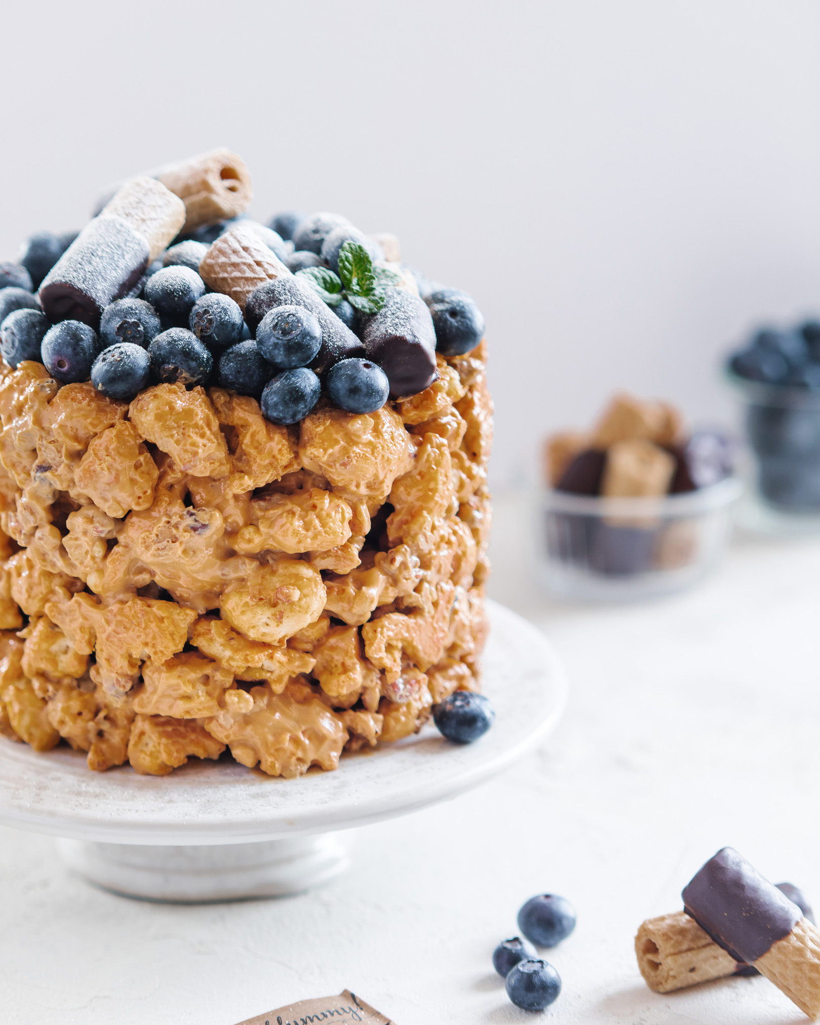 Cake with blueberries. A cake, topped with blueberries and mint, is in the middle of the photo. In the background, there are boxes with blueberries and cookies.