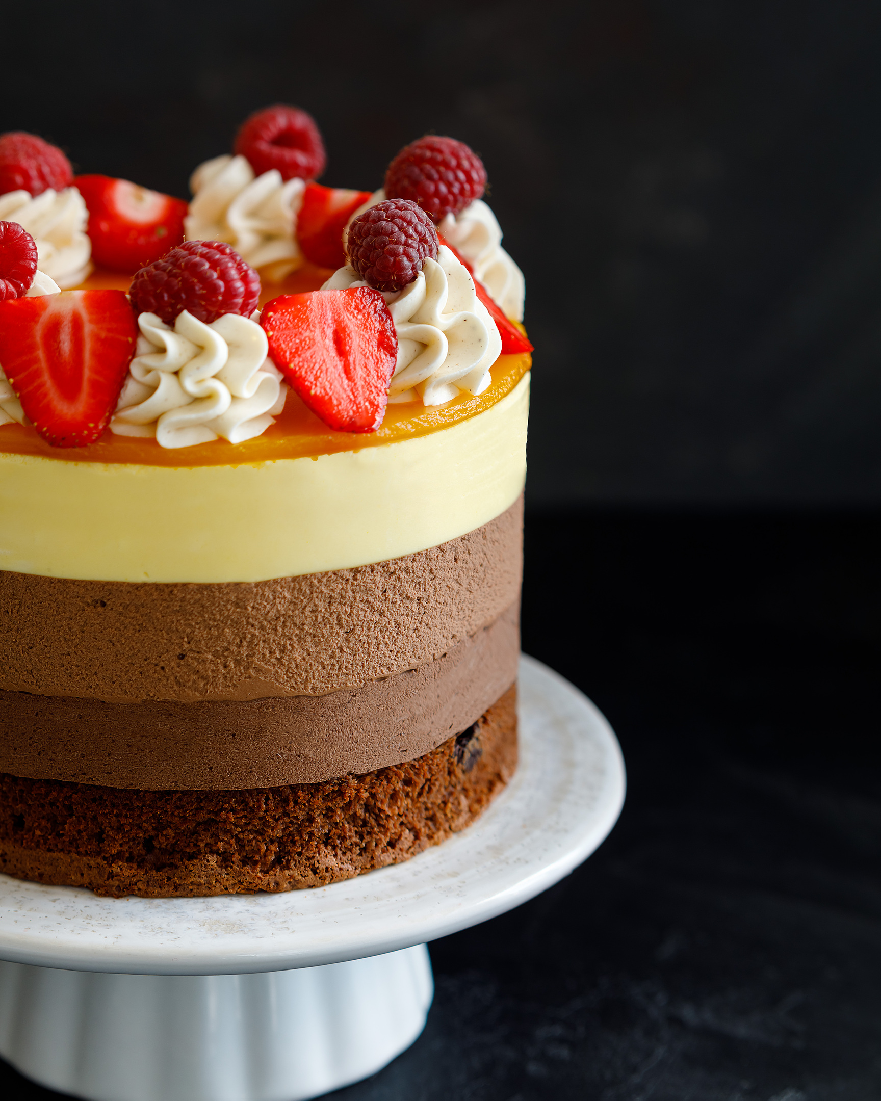 Шоколадова торта. В средата на снимката е шоколадова торта на постамент от бяла керамика. Върху него има ванилов крем, малини и ягоди. Можете да видите всеки слой от тортата!