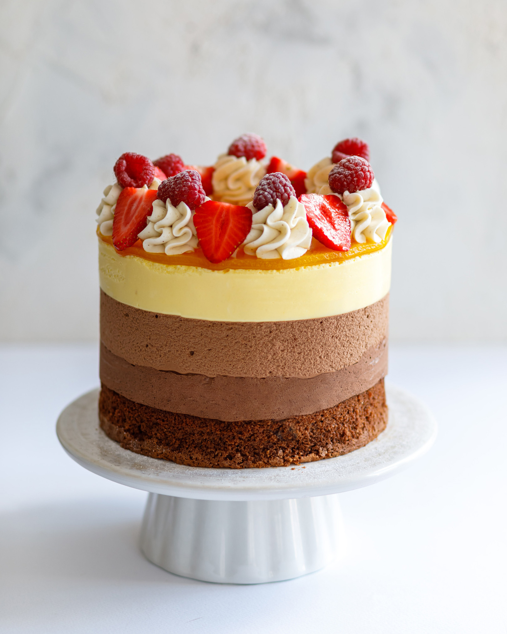 Chocolate Cake. In the middle of the photo is a chocolate cake on a pedestal made of white ceramics. On the top of it, there a vanilla cream, raspberries and strawberry. You can see every layer of the cake!