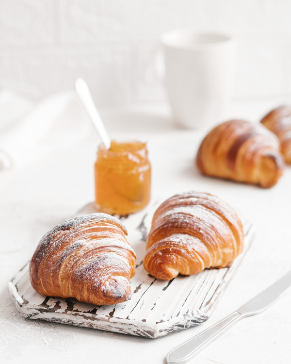 Croissants - Metro. Croissants - Metro. Two croissants are in the center of the photo, topped with sugar powder, on a craft desk. In the background, there are two more and a jar with an orange jam.