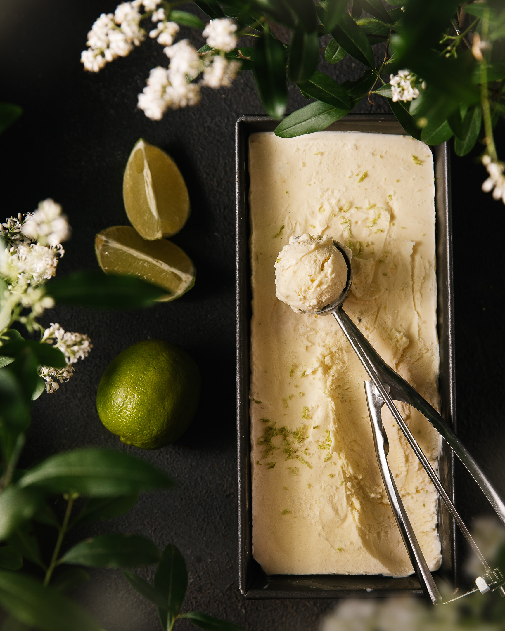 Lemon Ice Cream. A box with lemon ice cream is on the right in the dark background. There are flowers and leaves around. On the left side, there are green lemons. A special spoon for the ice cream is lying on the top of the box with the ice cream.