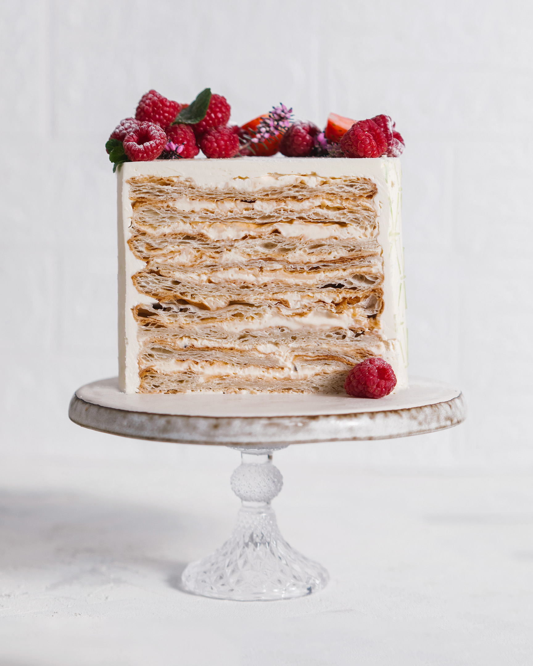 Napoleon Cake. In the middle of the photo, there is a Napoleon Cake on a pedestal made of glass. On the top of it, there are raspberry and mint. You can see every layer of the cake!