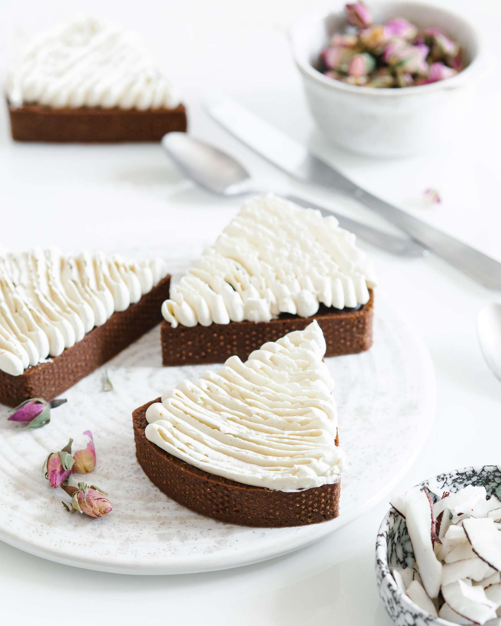 Pieces of a chocolate cake with vanilla cream and coconut. A few pieces of chocolate cake with vanilla cream are in the middle of the photo. There are bowls with roses and coconut on the right side.