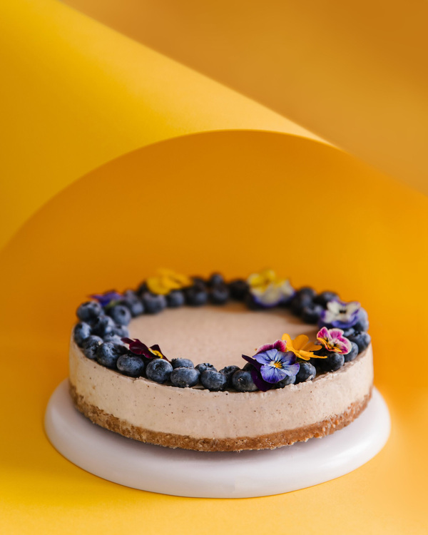 Round Cake With Blueberries. Round cake topped with blueberries and flowers on a pedestal made of white ceramic in the middle of the photo. A few layers of the cake are visible. The background is yellow and orange.