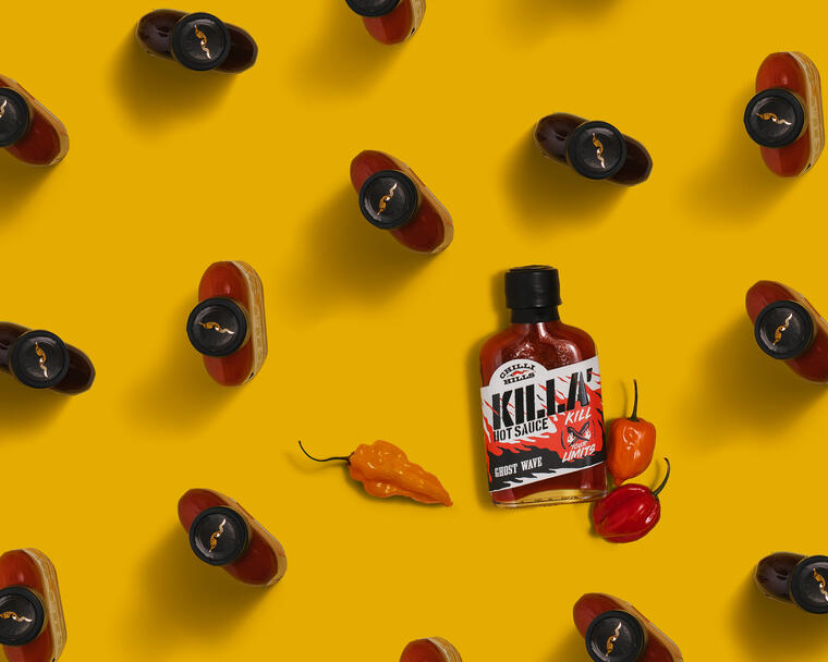 Сhilli Hills Killa Ghost Wave and another sauces from top view. On a yellow background lies a bottle of Chilli Hills Killa Ghost Wave. Near the bottle are habanero peppers, which demonstrate the sauce's main component. Throughout the entire picture area, there are bottles of the same manufacturer, but of different flavors, which are directed with their caps toward the viewer.