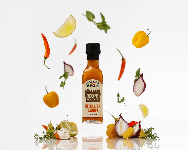 ChilliHills flying sauce Bulgarian Carrot. ChilliHills flying sauce Bulgarian Carrot. Against a light background, a jar of Chili Hills Bulgarian Carrot Sauce floats in the air. Ingredients that make up the sauce fly around the jar. Onion, pepper, spices, garlic. Vegetables lie on a light surface. The reflection of the jar and vegetables is visible, which informs the viewer that the objects are lying on a reflective surface.