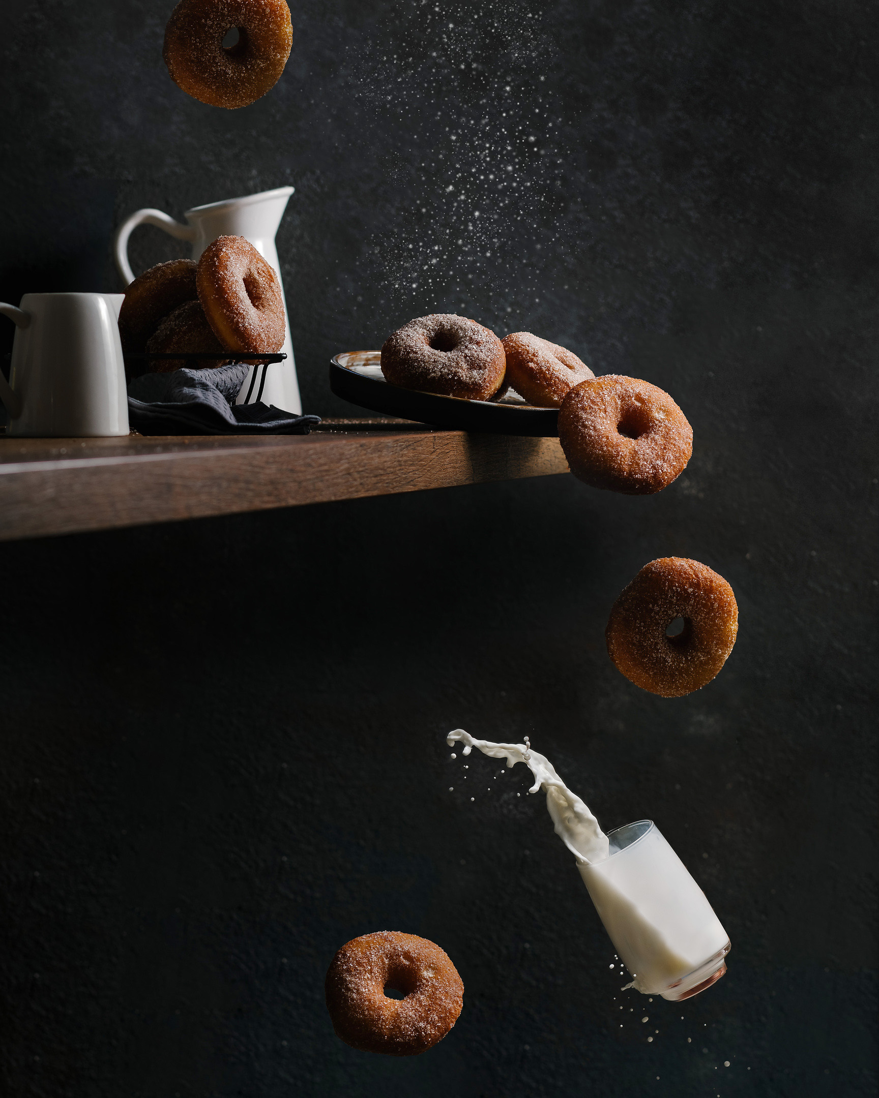 Flying Donuts And Milk Splash. On a dark background, the edge of the countertop is visible. There are several light-coloured milk jugs on the countertop. There is a plate on the edge of the tabletop. She is already tilted and is about to fall. There are several donuts on the plate,donuts covered in sugar crystals, but some donuts are already flying in the air and will soon fall completely. In the lower corner of the picture, a glass of milk is falling, which is already flying to the floor and will soon break. A splash of milk is already pouring out of the glass!