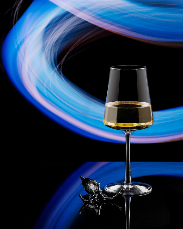 Glass of white wine with lights effects. Glass of white wine with lights effects. A glass of white wine stands on a dark background. Behind the goblet are visible ornate stripes of blue and violet hues. These same light effects are reflected in the surfaces under the glass. To the left of the glass lies a cork for a bottle of wine in the form of a silver stag's head