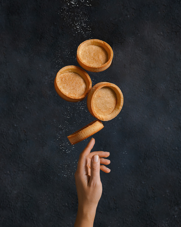 Empty tartlets hung in the air leaning on one finger of a female hand. An illusion demonstrating how tartlets hung in the air against a dark background, balancing and leaning on only one female finger. Additionally, the whole structure is sprinkled with powdered sugar.