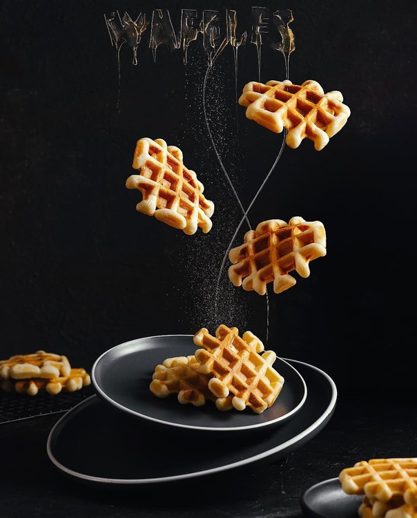 Waffles And Honey. Waffles And Honey. Plates stand on a dark background. They are tilted. On the top plate are some Viennese waffles. Honey pours thin strips on dashing. This honey flows from the inscription WAFFLE, which is also composed of honey. More waffles fly through the air. In the foreground and background of the photo, waffles just as simply lie. Powdered sugar is sprinkled in the center of the picture.